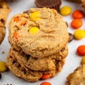 A stack of three peanut butter cookies with orange and yellow candy-coated chocolates, surrounded by more cookies and candies on a white surface.