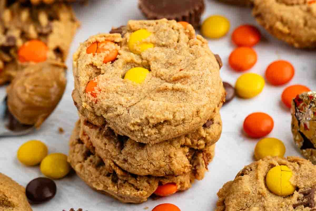 Pile de trois biscuits au beurre de cacahuète avec du chocolat enrobé de bonbons orange et jaune, entourés de plus de biscuits et de bonbons sur une surface blanche.