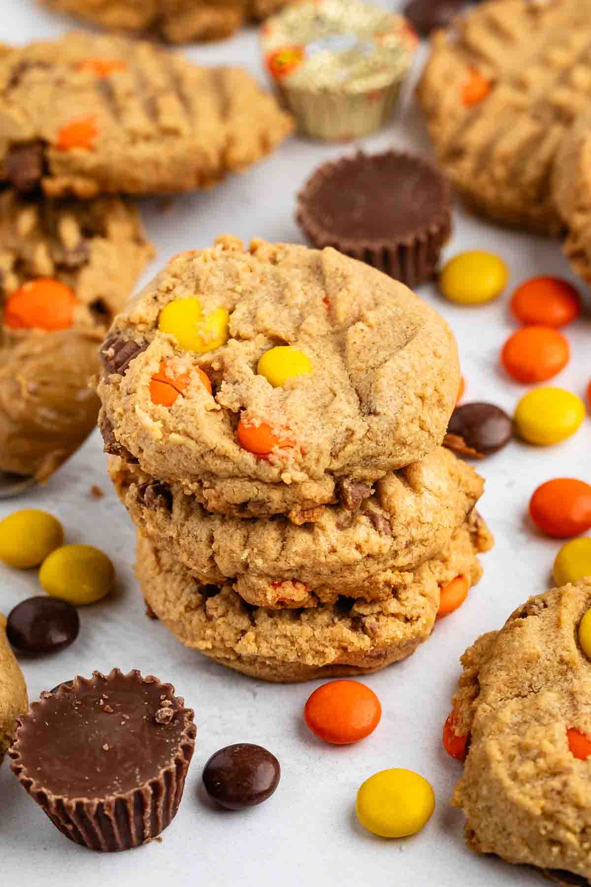 Une pile de biscuits au beurre de cacahuète avec des morceaux de bonbons colorés entourés d'autres biscuits, des coupes au beurre de cacahuète et des bonbons au chocolat dispersés sur une surface blanche.