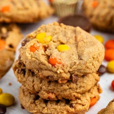 A stack of three thick, chewy peanut butter cookies with colorful Reese’s Pieces candies, surrounded by more candies, cookies, and mini peanut butter cups on a white surface.