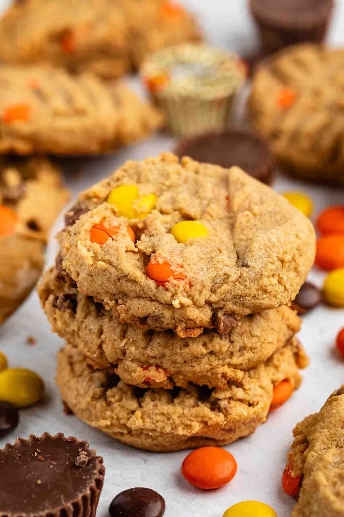 A stack of three thick, chewy peanut butter cookies with colorful Reese’s Pieces candies, surrounded by more candies, cookies, and mini peanut butter cups on a white surface.
