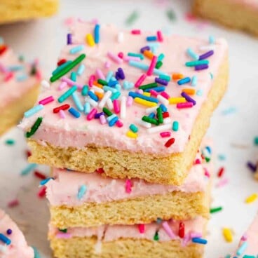 Three stacked sugar cookie bars topped with pink frosting and colorful rainbow sprinkles, with more cookie bars and scattered sprinkles in the background.