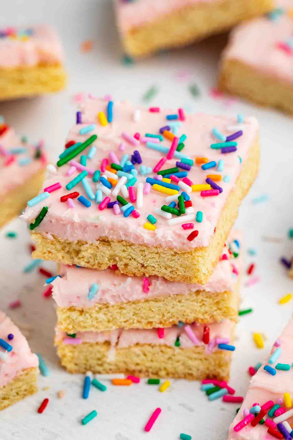 Meilleures barres de biscuits au sucre – Fou de croûte 5 Trois planches à biscuits au sucre empilées et recouvertes de glaçage rose et de pépites arc-en-ciel, avec d'autres planches à biscuits au sucre et pépites dispersées en arrière-plan.