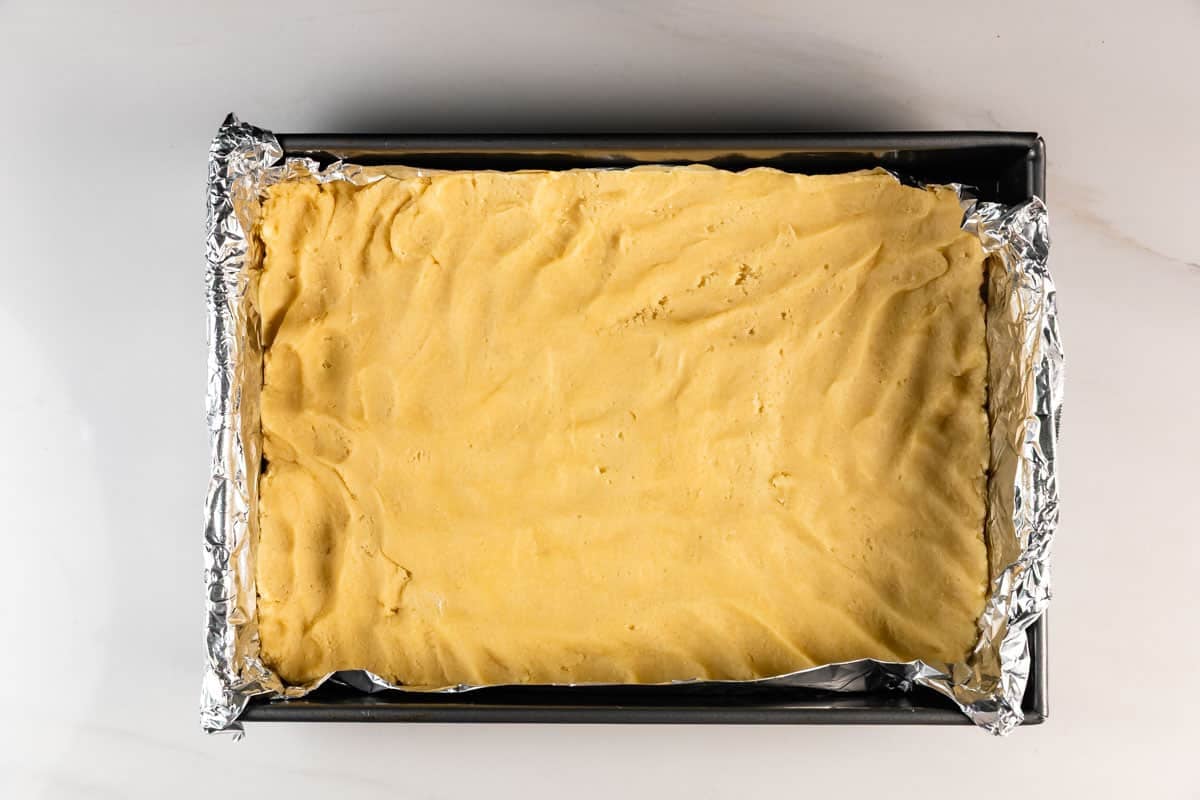 Meilleures barres de biscuits au sucre – Fou de croûte 12 Une plaque à pâtisserie rectangulaire est recouverte de papier d'aluminium et remplie d'une couche uniforme de pâte non cuite, et placée sur une surface blanche.