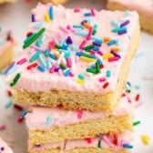 A stack of three sugar cookie bars topped with pink frosting and colorful rainbow sprinkles, with more cookie bars and sprinkles scattered in the background.