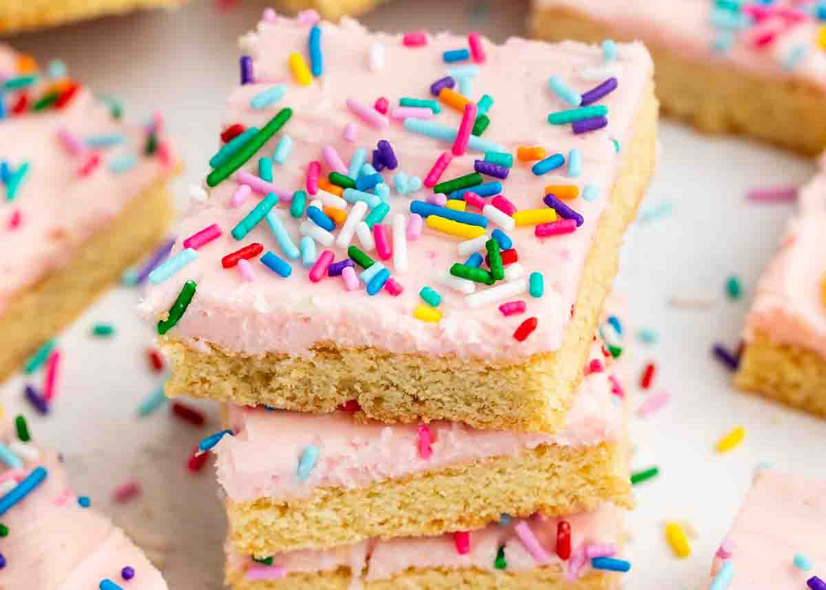 A stack of three sugar cookie bars topped with pink frosting and colorful rainbow sprinkles, with more cookie bars and sprinkles scattered in the background.
