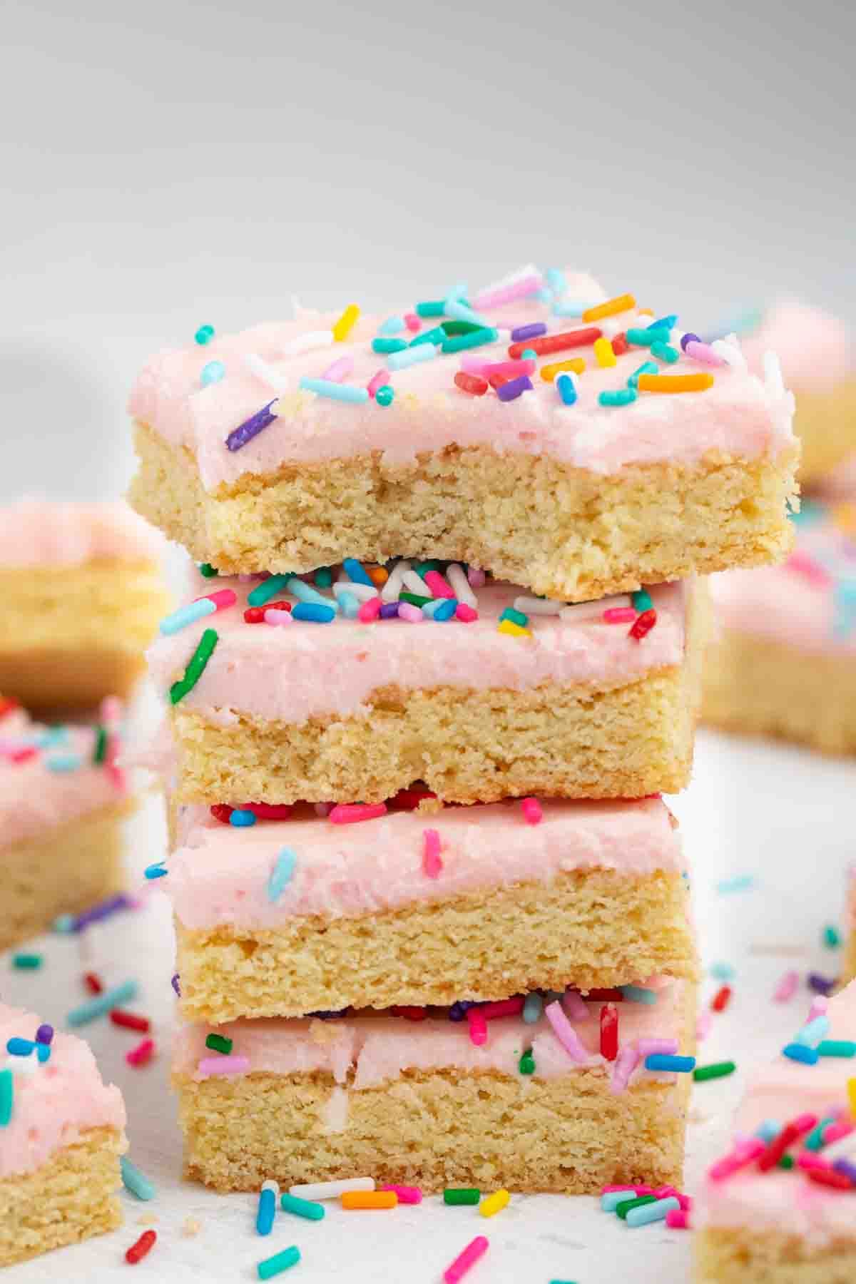 Meilleures barres de biscuits au sucre – Fou de croûte 7 Une pile de barres à biscuits au sucre givré avec un glaçage rose et des pépites arc-en-ciel, avec d'autres plaques à biscuits et des pépites éparpillées sur une surface blanche.
