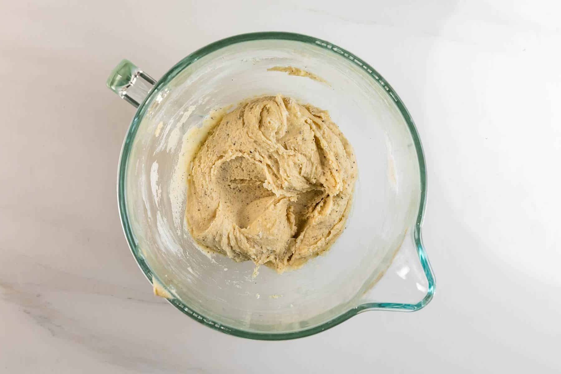 A glass mixing bowl filled with creamy, light brown cookie dough sits on a white marble surface.
