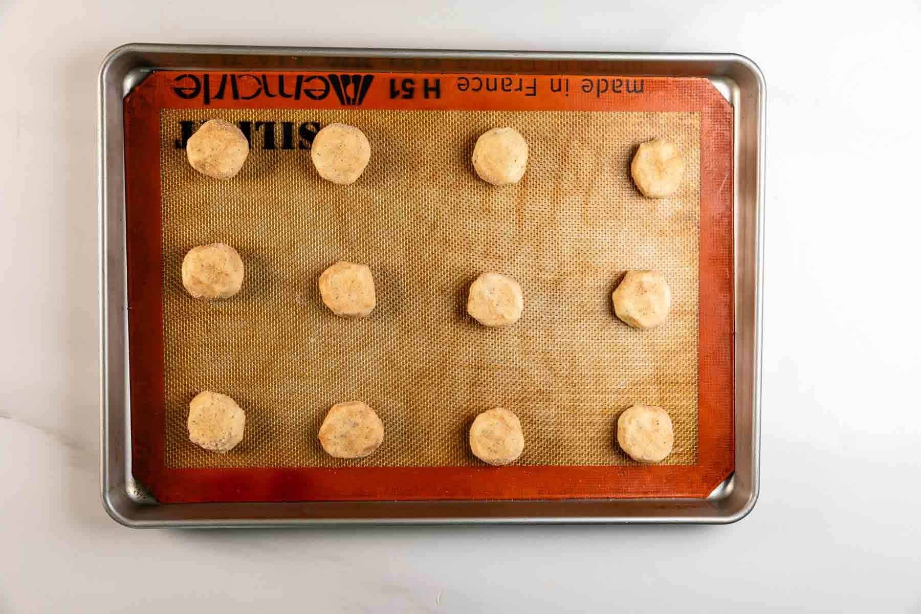 A baking tray lined with a silicone mat holds 12 evenly spaced balls of cookie dough, ready to be baked. The tray is placed on a white surface.