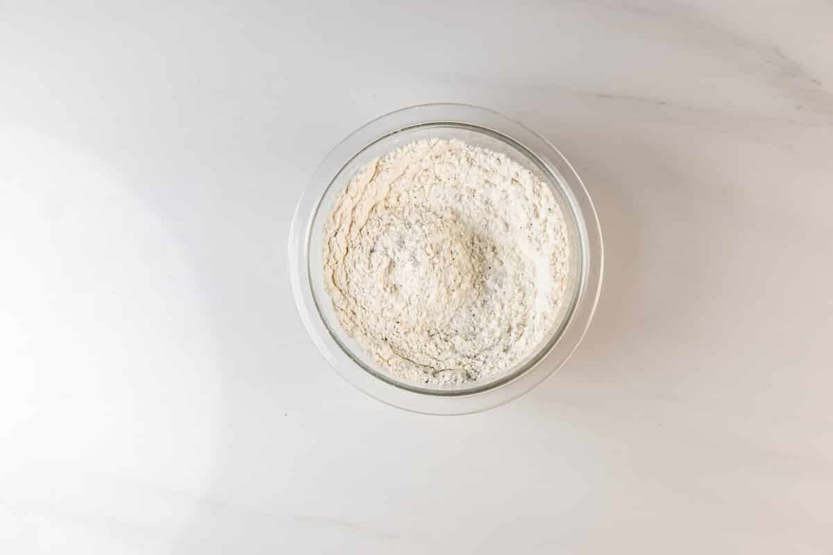 A glass bowl filled with white flour sits on a white marble surface, viewed from above.