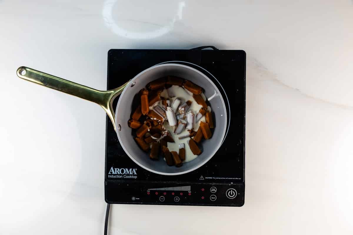 A saucepan with caramel candies, cream, and butter sits on an Aroma induction cooktop, viewed from above on a white countertop.
