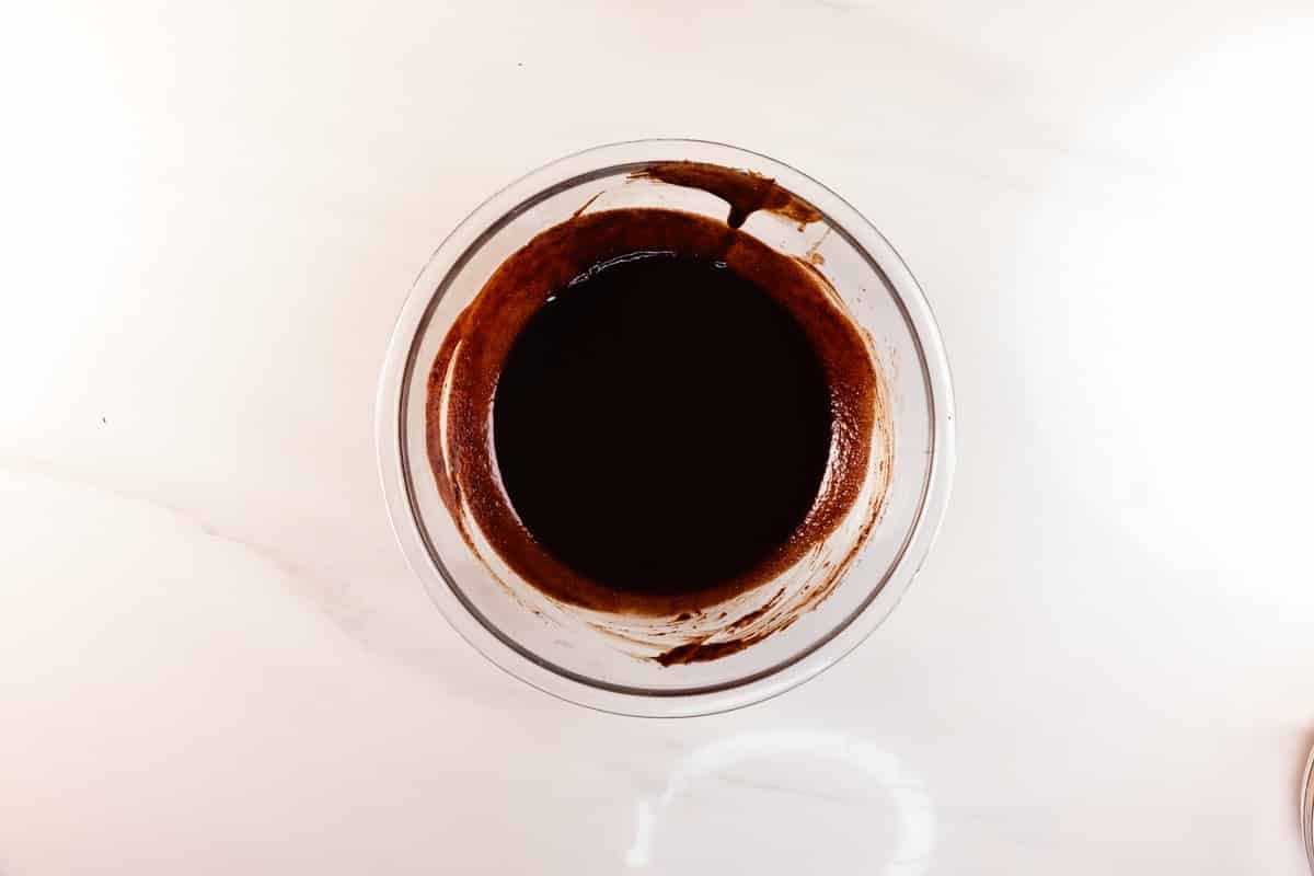 A glass bowl filled with dark chocolate batter sits on a white marble surface, viewed from above.