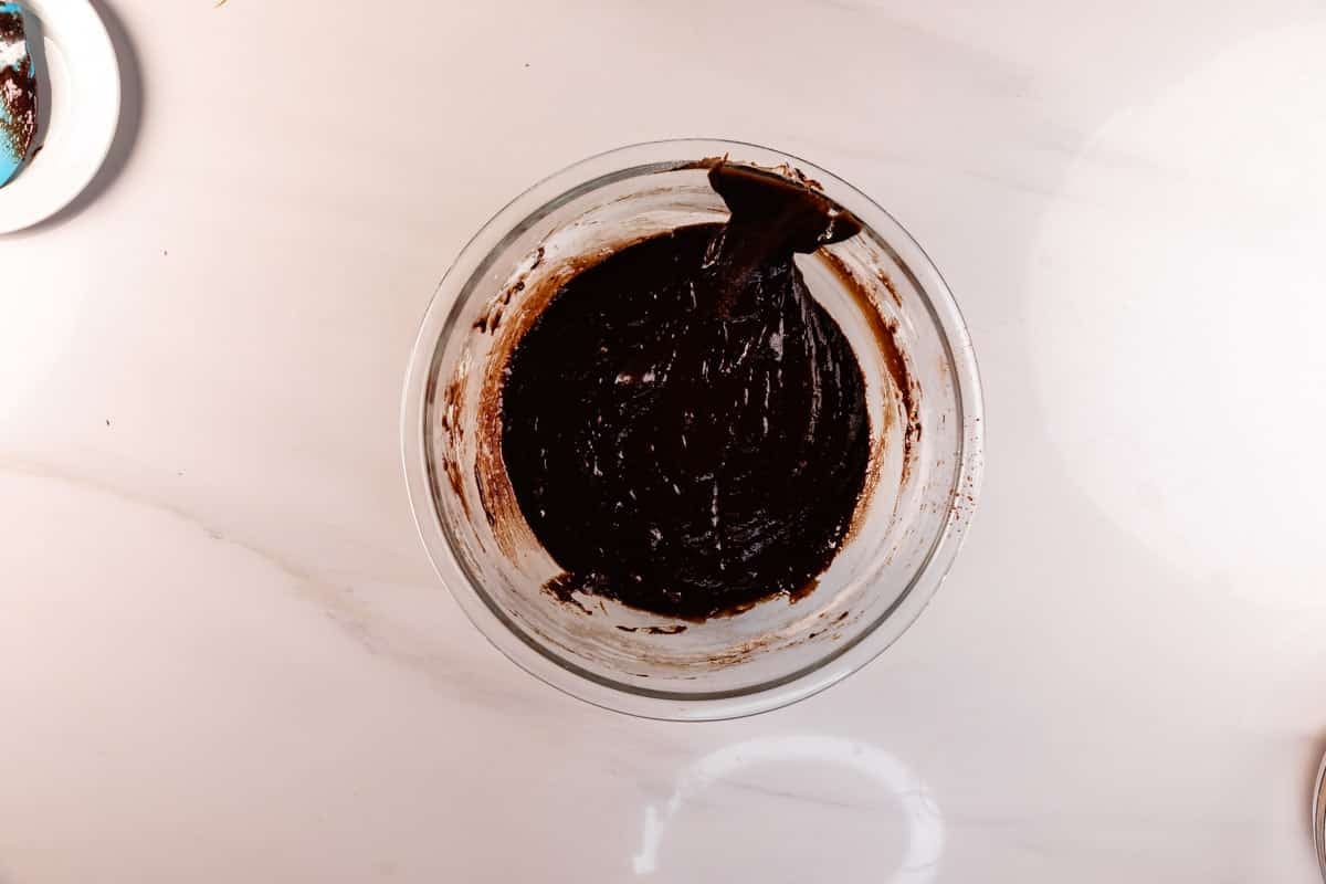 A glass bowl filled with thick chocolate batter sits on a white countertop, with a spatula resting on the edge of the bowl.