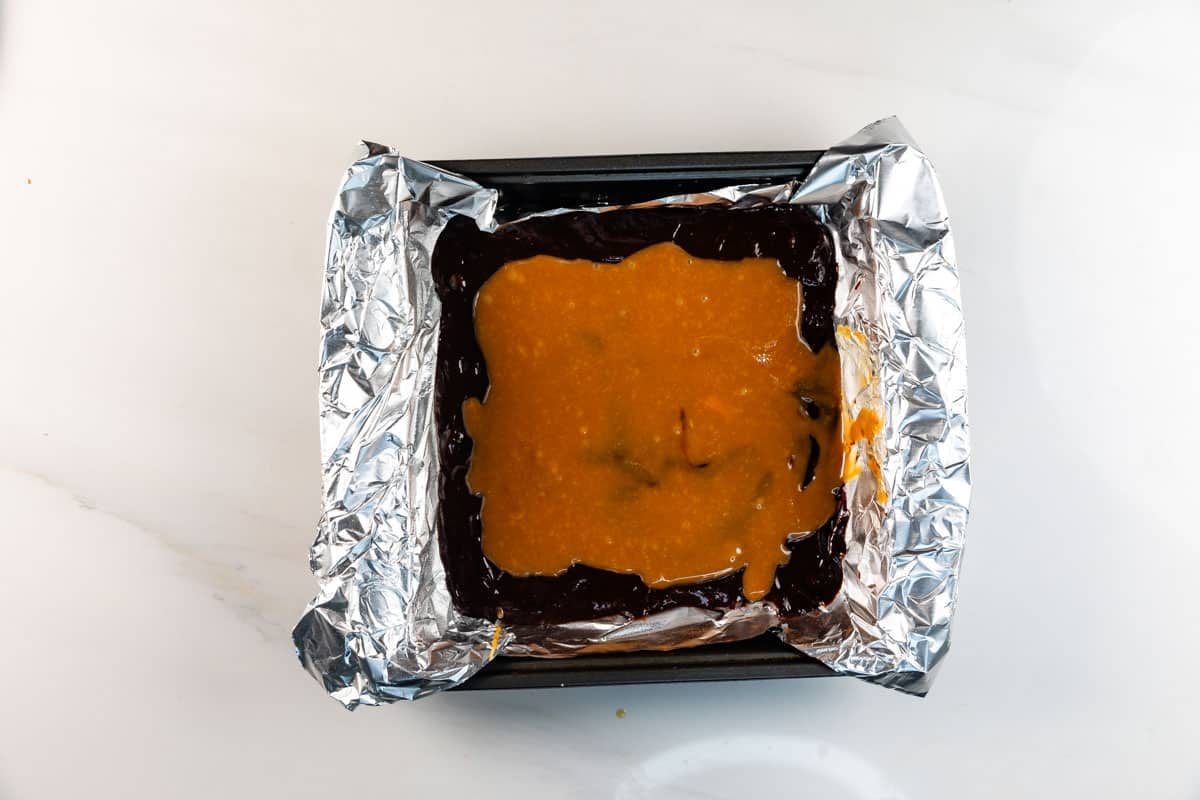 A square baking pan lined with crumpled aluminum foil, filled with a dark chocolate layer topped with a spread of orange-colored mixture, sits on a white surface.