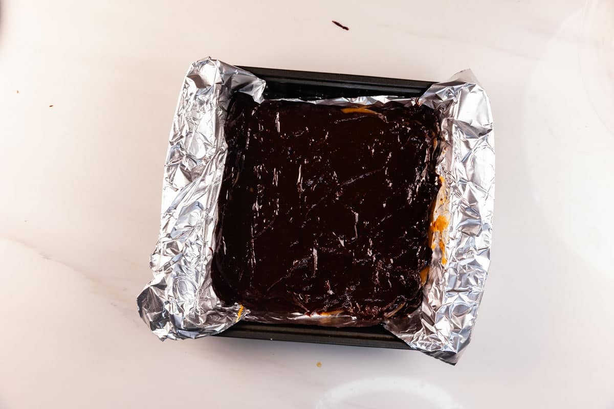 A square baking pan lined with crinkled aluminum foil, filled with a dark, glossy chocolate layer on top of a dessert, sits on a white surface.
