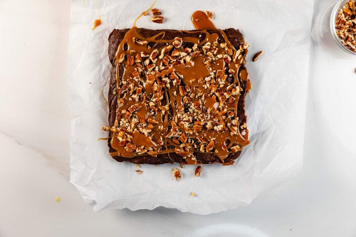 A square chocolate dessert topped with chopped pecans and caramel sauce sits on parchment paper on a white surface. Some pecan pieces and caramel are scattered around the dessert.