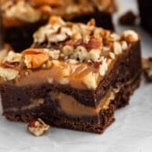 A close-up of a rich chocolate brownie topped with gooey caramel and chopped pecans, with more brownies and caramel pieces in the background on a white surface.