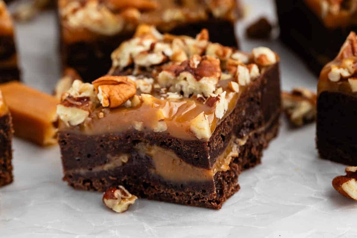 A close-up of a rich chocolate brownie topped with gooey caramel and chopped pecans, with more brownies and caramel pieces in the background on a white surface.