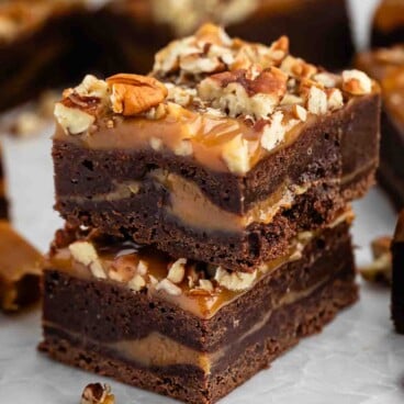 Two stacked chocolate brownies with caramel and chopped pecans on top sit on parchment paper, with more brownies and pecan pieces in the background.