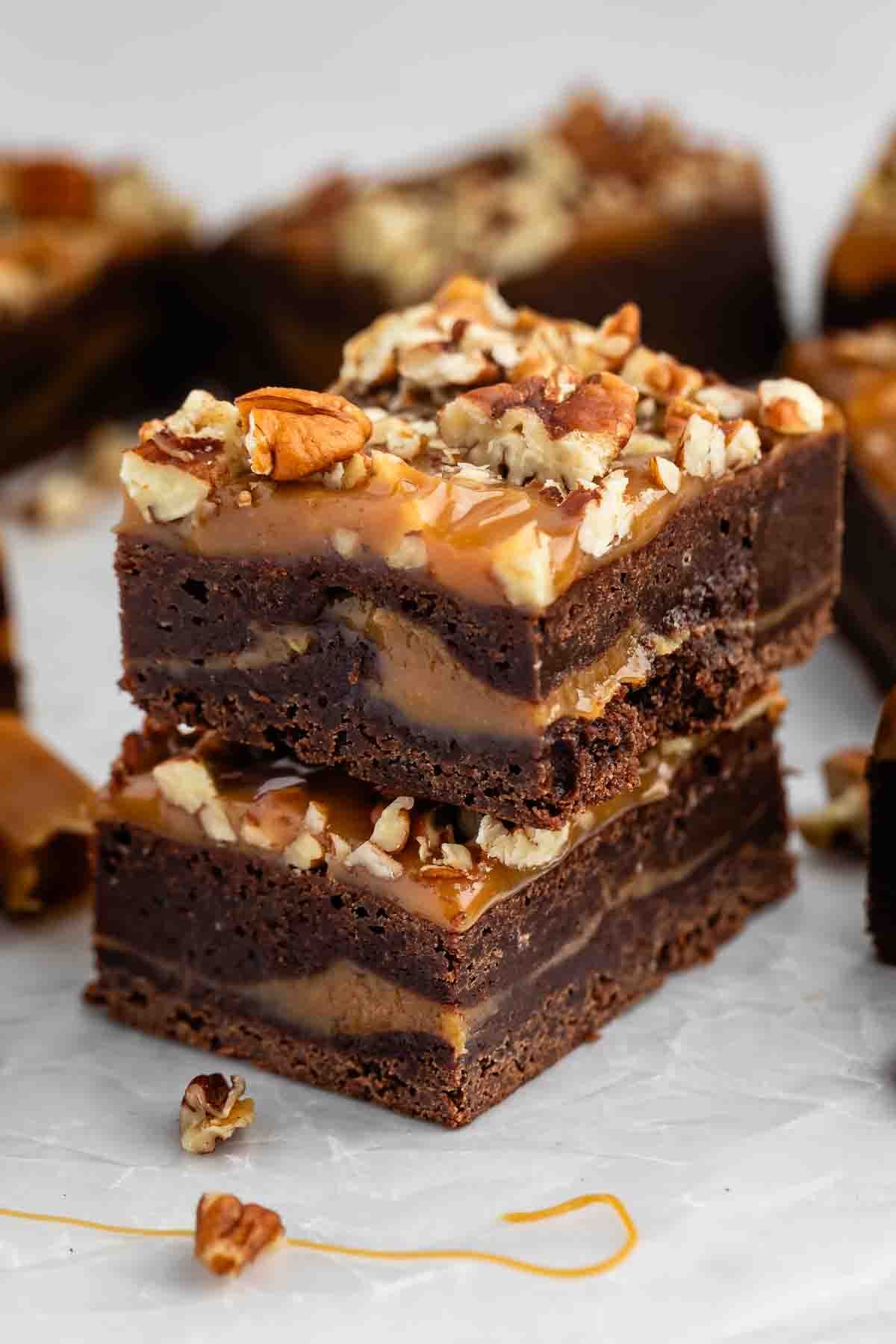 Two stacked chocolate brownies with caramel and chopped pecans on top sit on parchment paper, with more brownies and pecan pieces in the background.