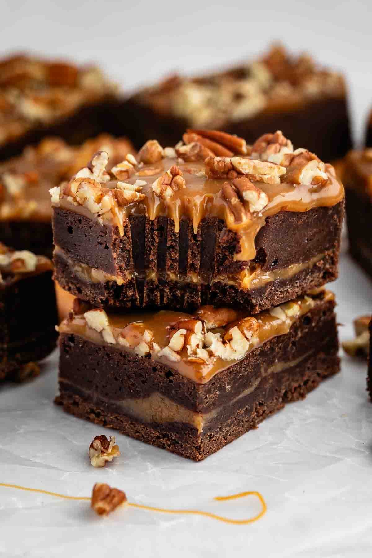 Two stacked caramel pecan brownies topped with chopped pecans and drizzled caramel sauce, with more brownies and pecan pieces scattered in the background on a white surface.
