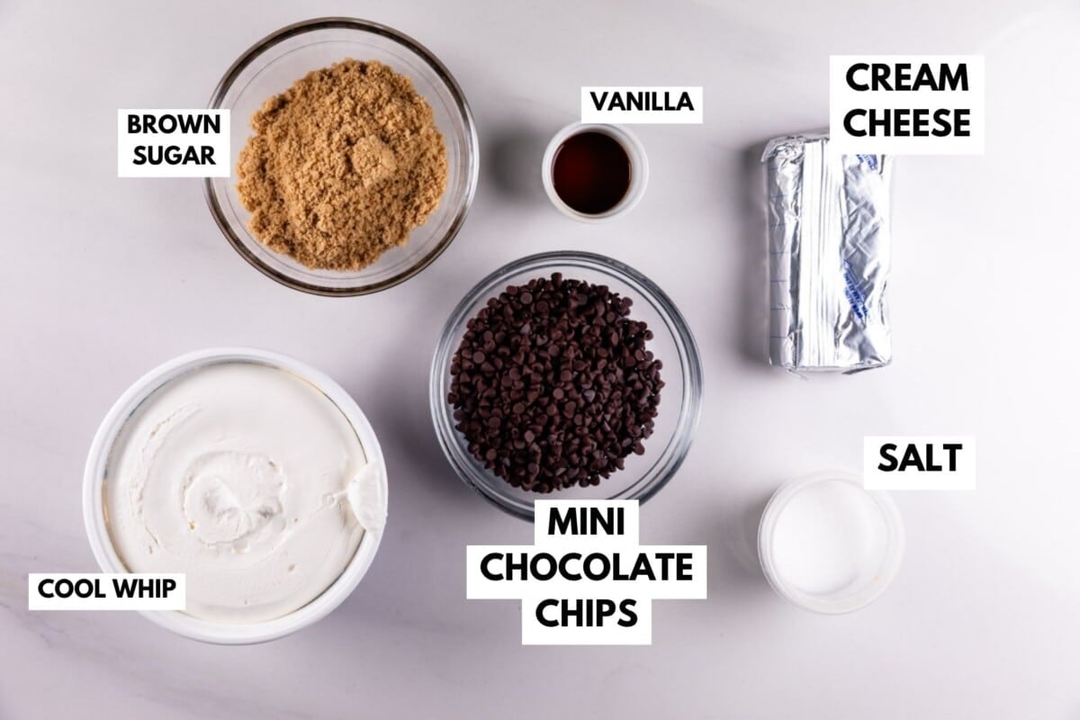 Top-down view of labeled baking ingredients on a white surface: brown sugar, vanilla, cream cheese, salt, mini chocolate chips, and cool whip in separate bowls or containers.