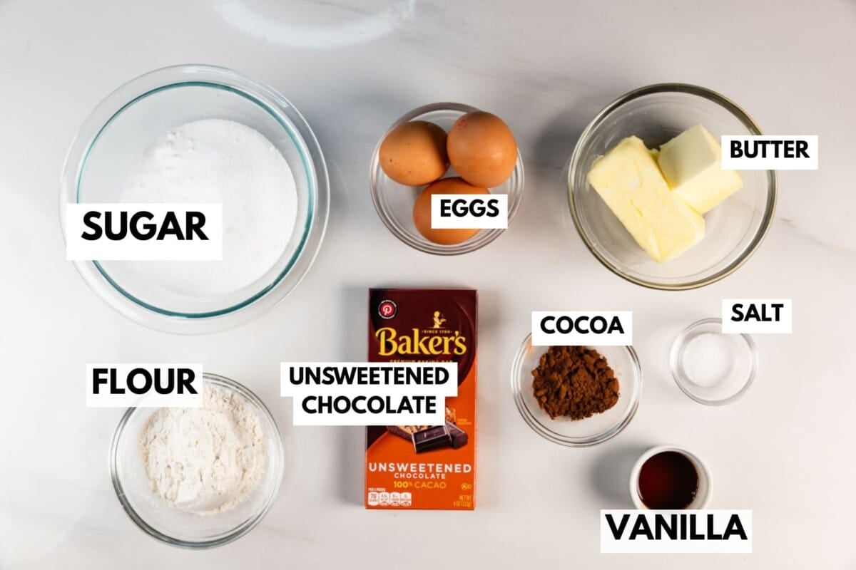 Une vue de haut en bas des ingrédients de cuisson dans les bols : sucre, farine, œufs, beurre, chocolat non sucré, poudre de cacao, sel et extrait de vanille, chacun étant étiqueté en gras.
