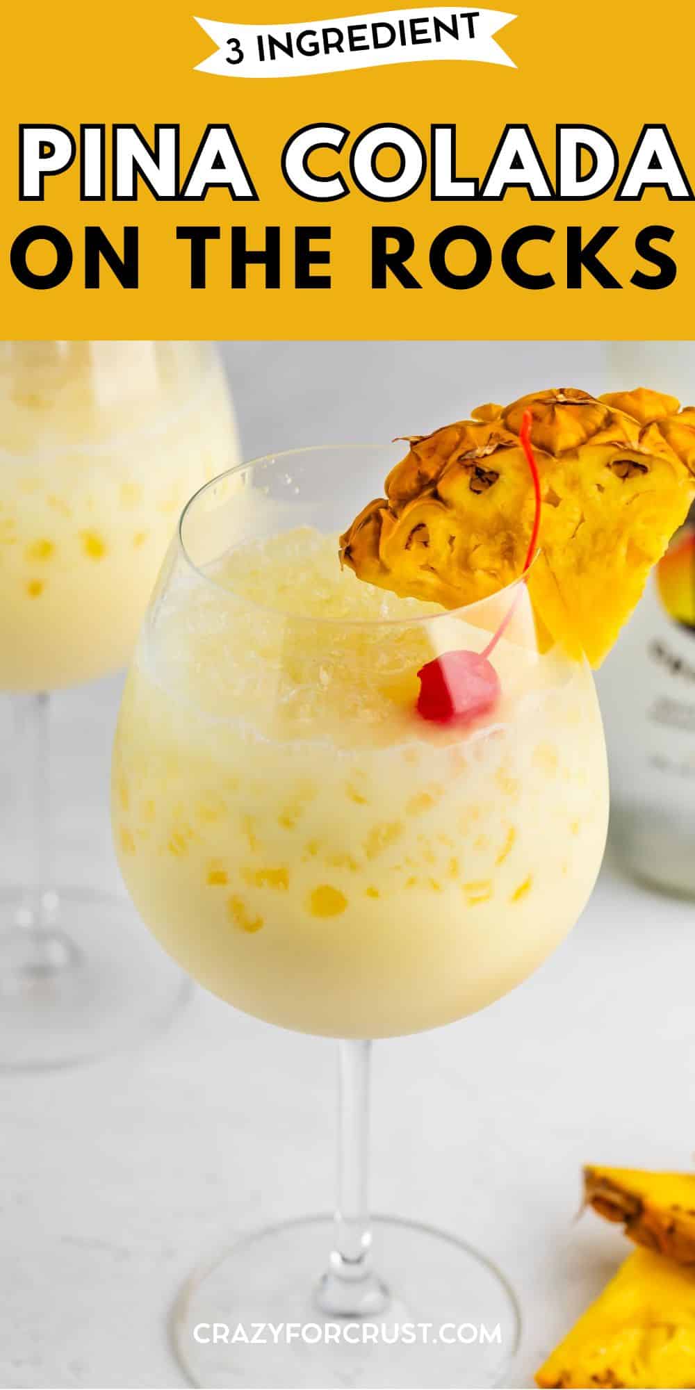 A pina colada served on the rocks in a wine glass, garnished with a pineapple slice and a maraschino cherry, with a second glass and pineapple pieces in the background. Text reads “3 Ingredient Pina Colada on the Rocks.”.