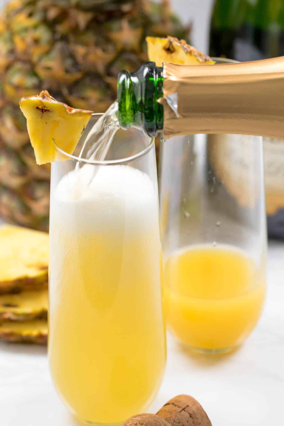 Champagne being poured into a flute glass with pineapple juice, garnished with a pineapple wedge; a fresh pineapple and juice are in the background.