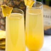 Two champagne flutes filled with yellow pineapple mimosa cocktails, garnished with pineapple wedges. A pineapple, champagne bottle, and cork are in the background.