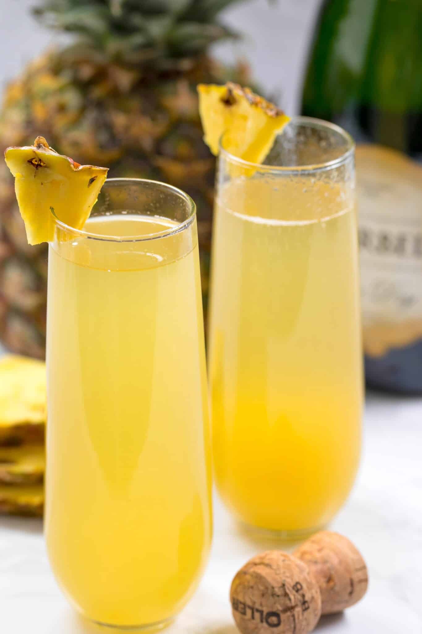 Two champagne flutes filled with yellow pineapple mimosa cocktails, garnished with pineapple wedges. A pineapple, champagne bottle, and cork are in the background.