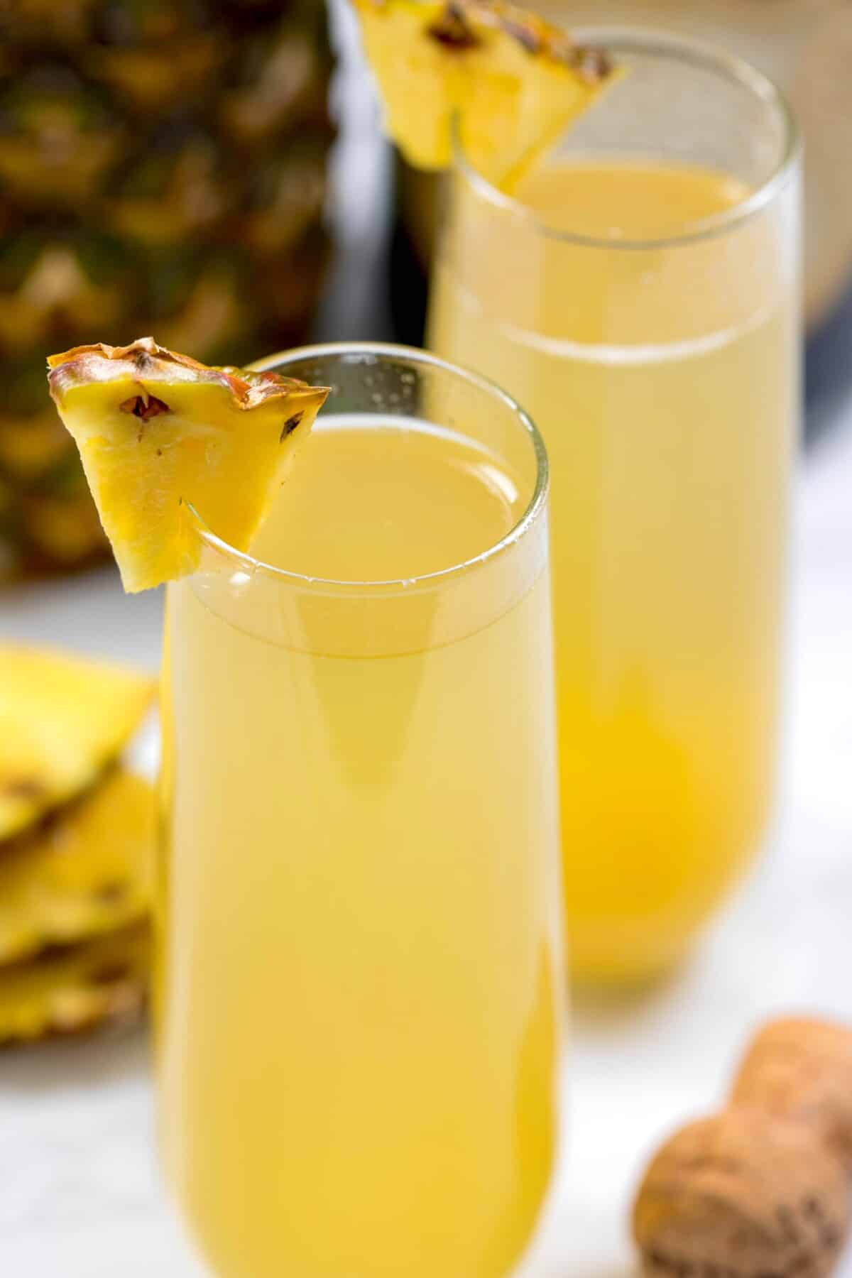 Two tall glasses filled with a yellow tropical drink, each garnished with a pineapple wedge. There are additional pineapple slices and a whole pineapple blurred in the background.