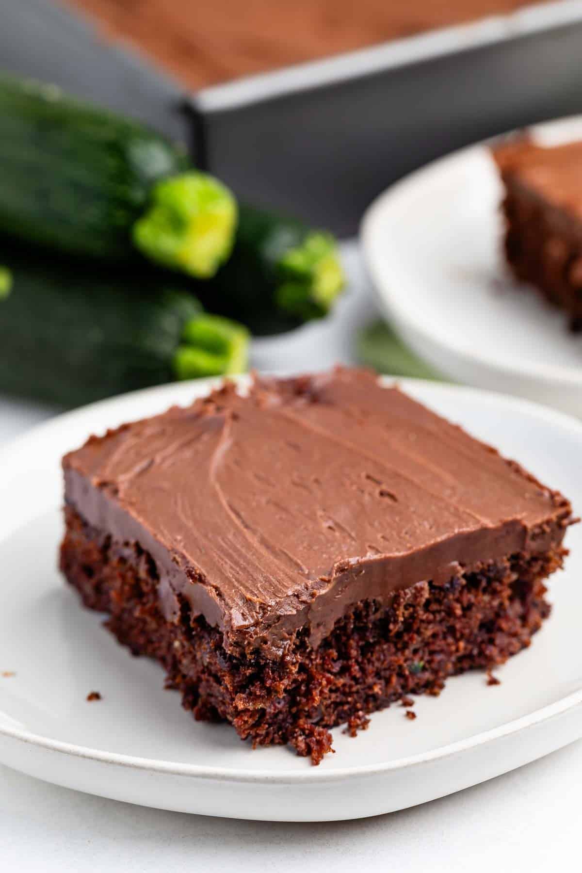 A slice of chocolate cake with chocolate frosting sits on a white plate. In the background, whole zucchinis and a baking tray with more frosted cake are visible.