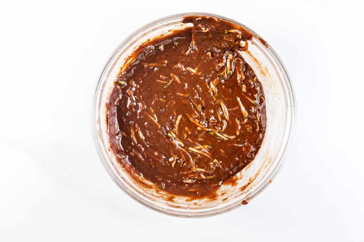 A clear glass bowl filled with a thick, chocolate-colored batter, possibly containing shredded zucchini, sits on a white surface.