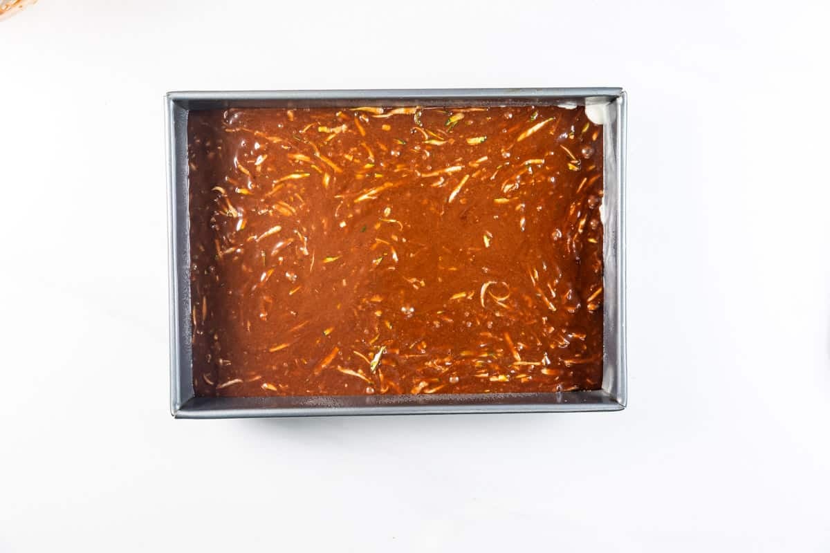 A rectangular metal baking pan filled with a brown, textured batter, possibly for a cake or brownies, sits on a white surface.
