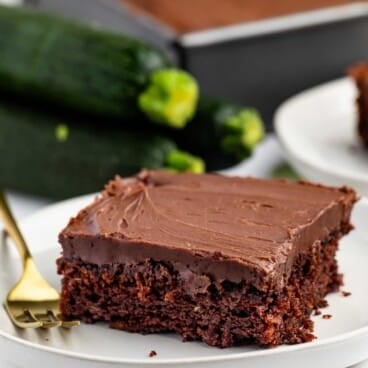 A slice of chocolate cake with chocolate frosting on a white plate, with a gold fork beside it. In the background, whole zucchinis and a tray of additional cake are visible.