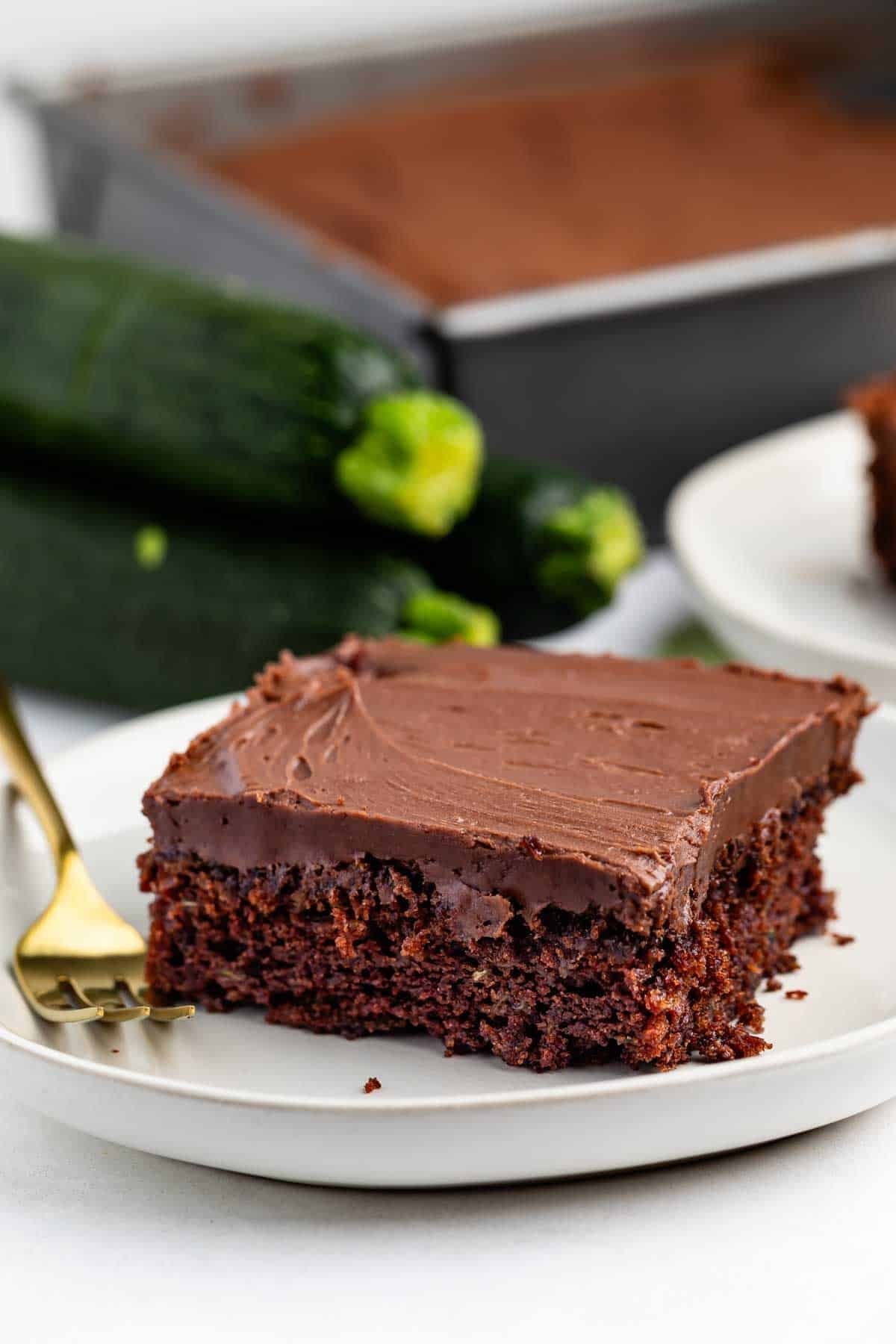 A slice of chocolate cake with chocolate frosting on a white plate, with a gold fork beside it. In the background, whole zucchinis and a tray of additional cake are visible.