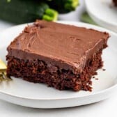 A square slice of chocolate cake with chocolate frosting sits on a white plate next to a gold fork, with whole zucchinis blurred in the background.