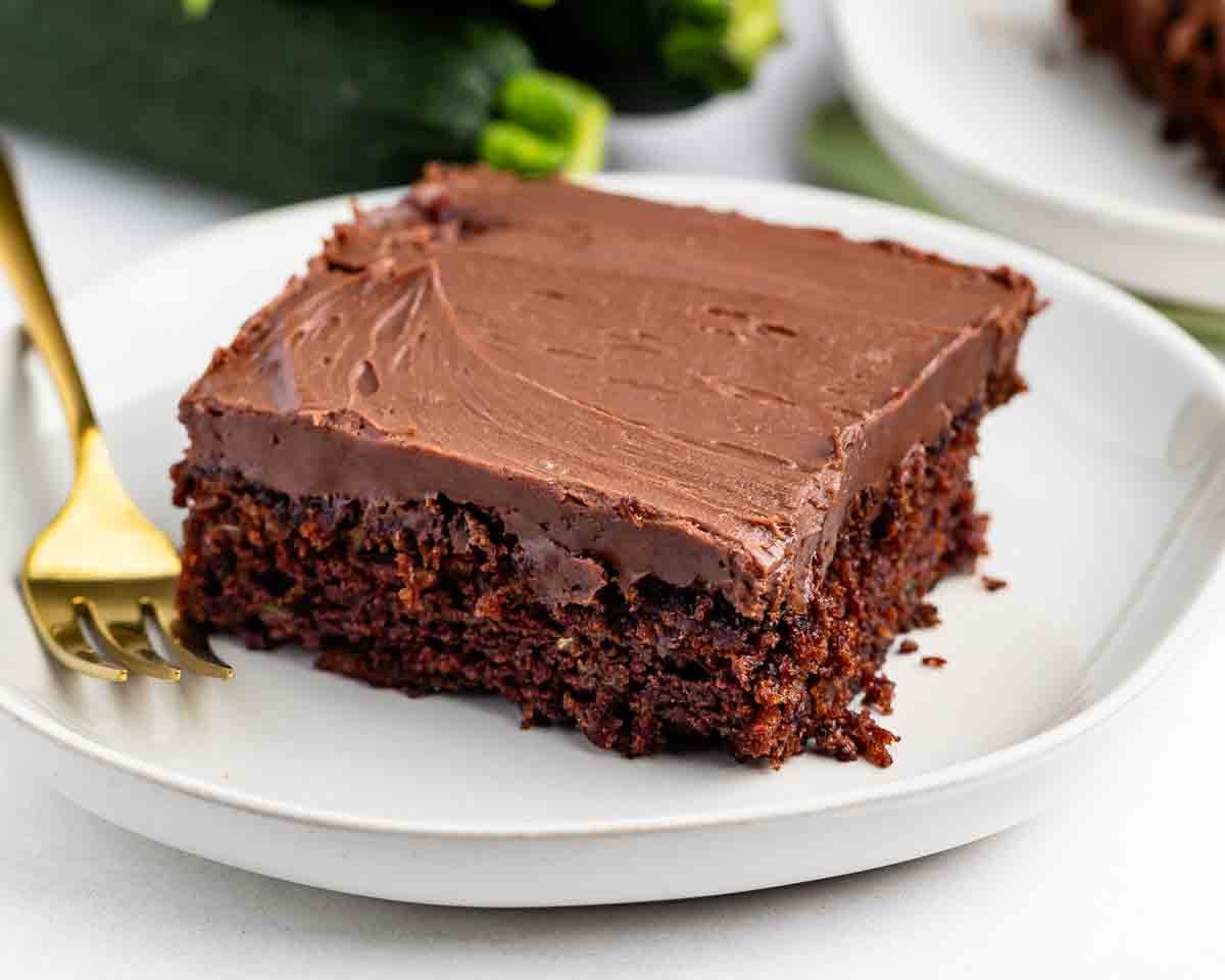 A square slice of chocolate cake with chocolate frosting sits on a white plate next to a gold fork, with whole zucchinis blurred in the background.