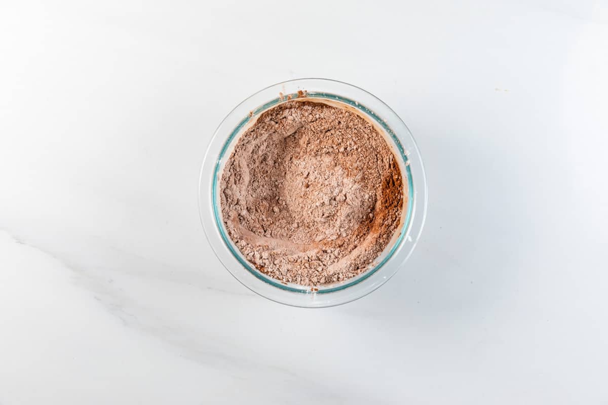 A glass bowl filled with cocoa powder and dry baking mix sits on a white marble surface.