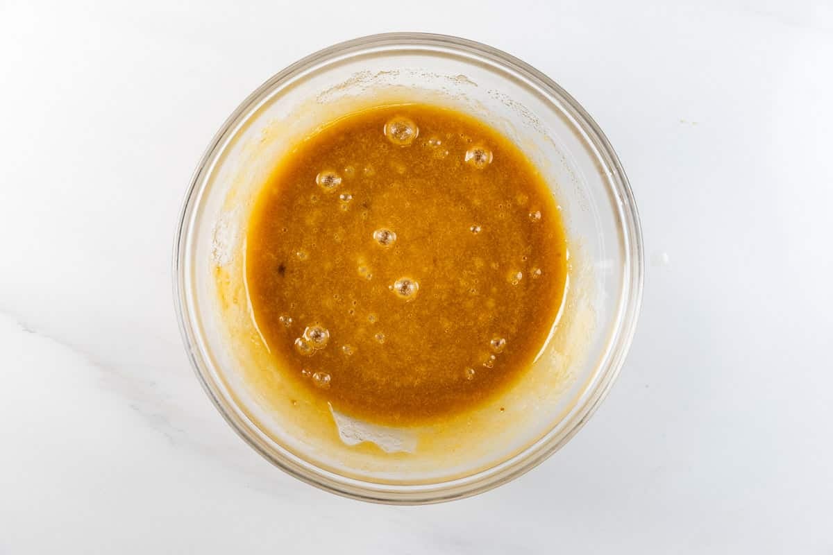 A clear glass bowl filled with a light brown, slightly frothy liquid mixture sits on a white surface.