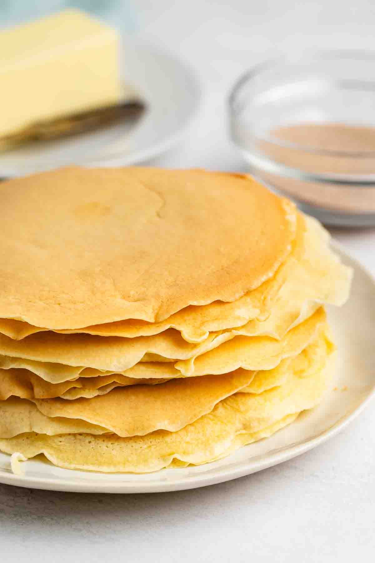 Rouleaux de tarte au sucre et à la cannelle - Fous de croûte 8 Une pile de délicates crêpes dorées posées sur une assiette blanche. En arrière-plan se trouve une assiette de beurre et un petit bol en verre rempli de poudre brune, peut-être de cannelle ou de sucre.