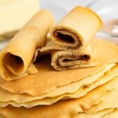 A stack of thin, golden-brown crepes with three rolled crepes placed on top, one of which is cut to show a light brown filling inside. Butter and a blurred utensil are in the background.