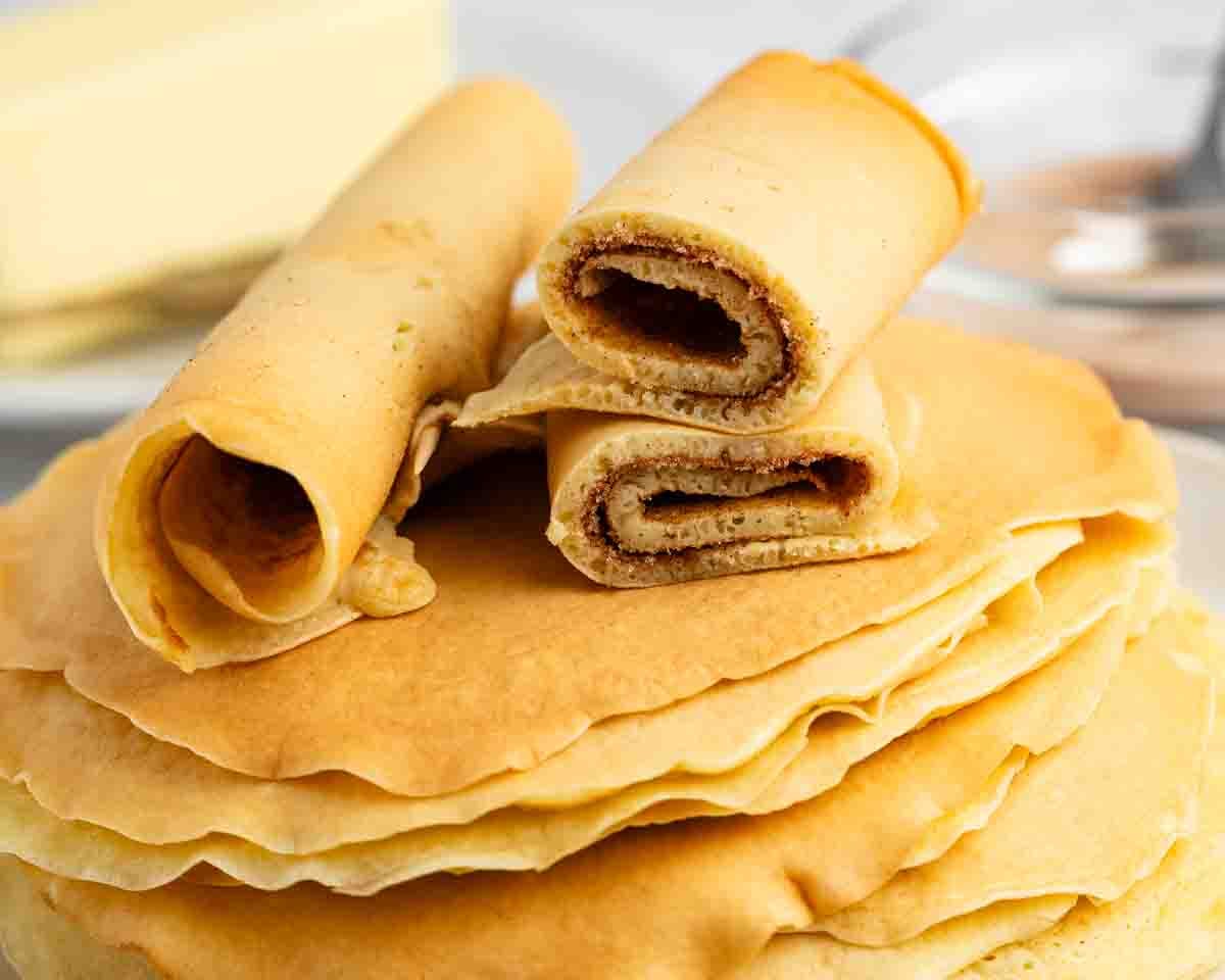 A stack of thin, golden-brown crepes with three rolled crepes placed on top, one of which is cut to show a light brown filling inside. Butter and a blurred utensil are in the background.