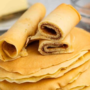 A stack of thin, golden Norwegian pancakes on a plate, with some pancakes rolled up and sliced to show a cinnamon filling. A stick of butter and a glass bowl are in the background.