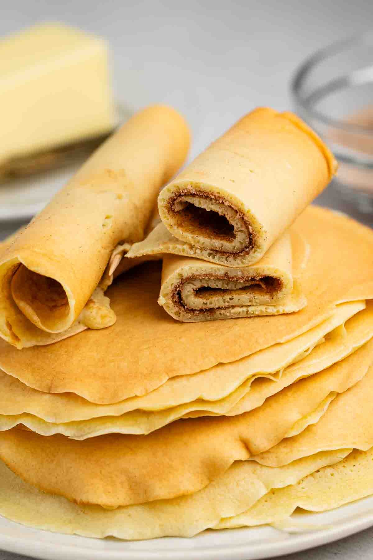 Rouleaux de tarte au sucre et à la cannelle - Fous de croûte 5 Une pile de crêpes norvégiennes moelleuses et dorées sur une assiette, avec certaines crêpes enroulées et coupées en tranches pour mettre en valeur la garniture à la cannelle. Il y a un bâton de beurre et un bol en verre en arrière-plan.