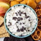 A bowl of creamy dip topped with chocolate chips is surrounded by apple slices, vanilla wafer cookies, pretzels, graham crackers, and chocolate sandwich cookies on a platter.