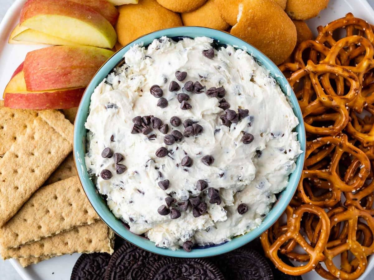 A bowl of creamy dip topped with chocolate chips is surrounded by apple slices, vanilla wafer cookies, pretzels, graham crackers, and chocolate sandwich cookies on a platter.