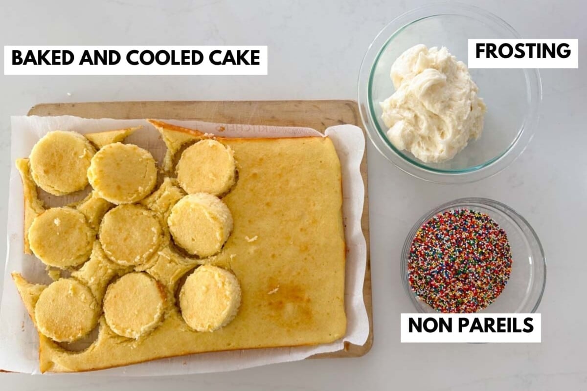 A rectangular baked cake with several circular pieces cut out sits on a wooden board beside bowls of white frosting and multicolored nonpareil sprinkles, each labeled accordingly.