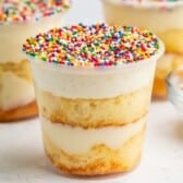 A clear plastic cup filled with layers of vanilla cake and creamy frosting, topped with colorful rainbow sprinkles. Two similar cups and a glass bowl of sprinkles are in the background.